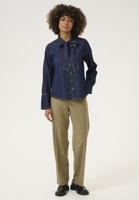 Blusa in denim di colore blu scuro con colletto a fiocco, chiusura frontale con bottoni e maniche lunghe. Abbinata a pantaloni beige e scarpe nere.
