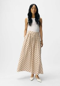 Falda maxi de lunares beige con pliegues, bolsillos laterales, un ajuste relajado y combinada con una camiseta ajustada blanca y zapatos de punta blancos.