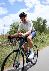 Vélo de route léger bleu avec des guidons plongeants et des roues noires. Le cycliste porte un maillot blanc à manches courtes et un cuissard de cyclisme noir.