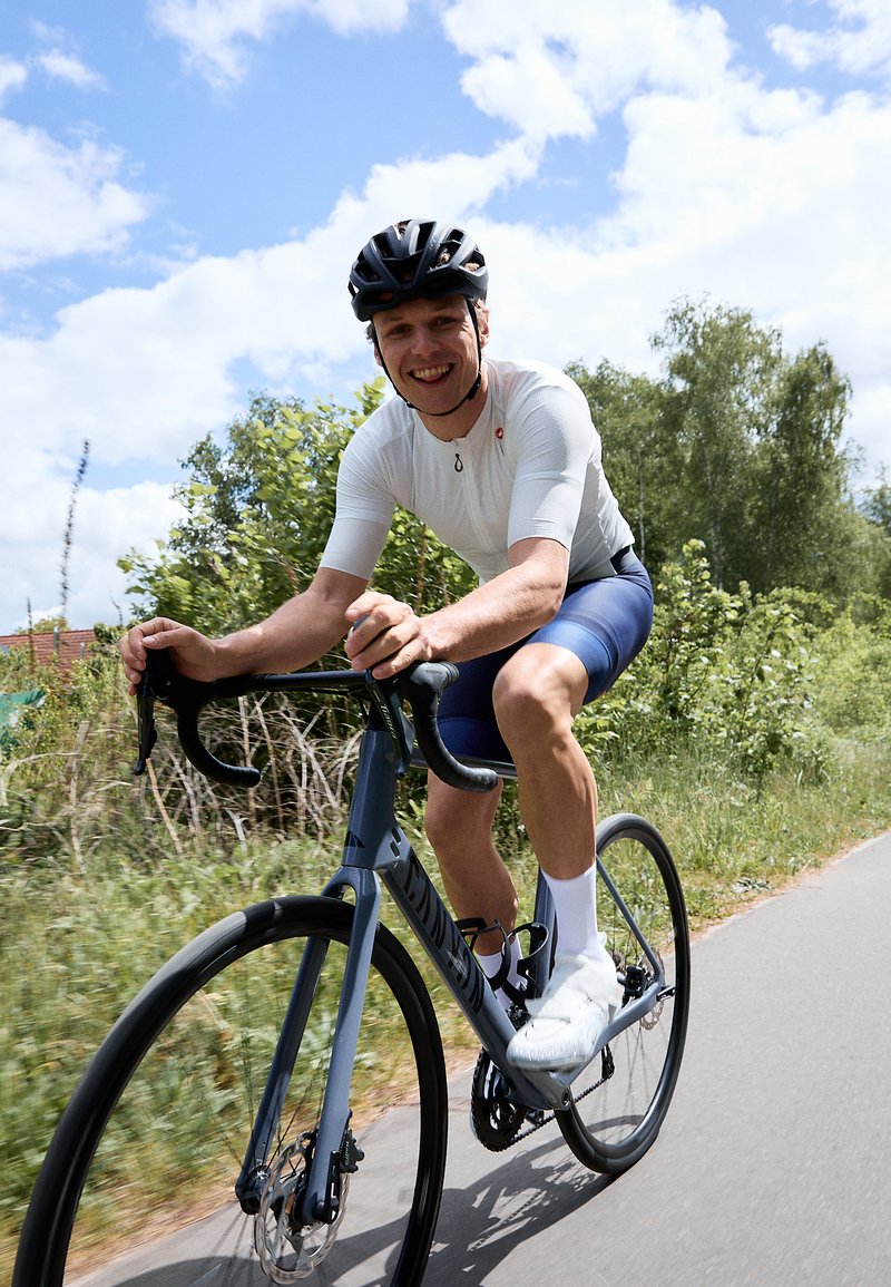 Vélo de route léger bleu avec des guidons plongeants et des roues noires. Le cycliste porte un maillot blanc à manches courtes et un cuissard de cyclisme noir.