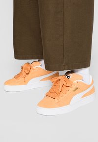 Orange suede sneakers with white accents, featuring thick laces, a textured sole, and a gold logo on the side.