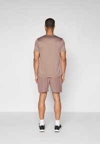 Manns sporty antrekk som inkluderer en kortermet, lys brun skjorte og matchende shorts, begge i et glatt og lett stoff. Svarte joggesko fullfører stilen.