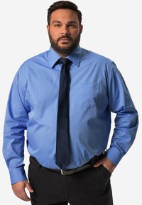 Camicia azzurro chiaro con colletto classico, maniche lunghe, taschino sul petto e cravatta scura, abbinata a pantaloni neri e una cintura.