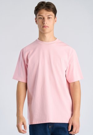 Junger Mann mit kurzem braunem Haar, der ein schlichtes, hellrosa übergroßes T-Shirt und blaue Jeans trägt, vor einem hellen Hintergrund stehend.