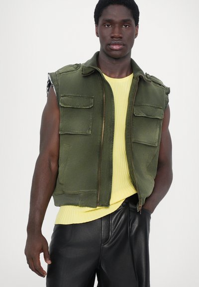 Gilet utilitaire vert avec fermeture éclair à l'avant et quatre poches, superposé à un t-shirt jaune côtelé, associé à un pantalon en cuir noir.