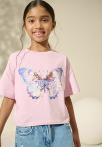 Camiseta de algodón rosa de manga corta con un gráfico de mariposa colorido y acentos de purpurina. Combinada con pantalones cortos de mezclilla.