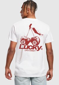 Fehér pamut póló, amelyen egy grafika látható két cseresznyével, a "Lucky 8" és a "LIFE'S SWEET" felirattal piros színben. Normál fazon, rövid ujjú.