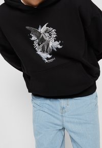 Černá mikina s kapucí s grafikou anděla uprostřed obláčkových vzorů. Design obsahuje text a kontrastuje s světle modrými džínami.