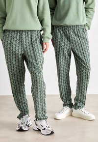 Duas pessoas vestindo calças verdes com padrão, sweatshirts verdes claras e tênis brancos, de pé lado a lado em um piso neutro.
