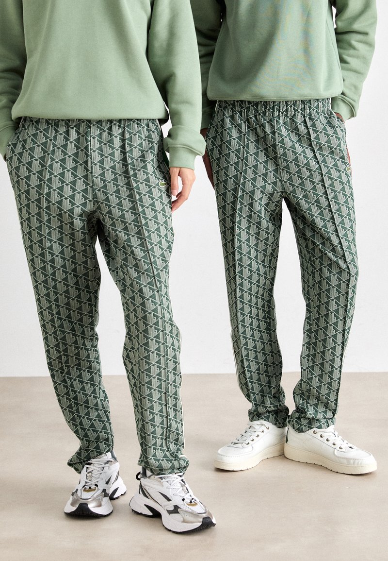 Duas pessoas vestindo calças verdes com padrão, sweatshirts verdes claras e tênis brancos, de pé lado a lado em um piso neutro.