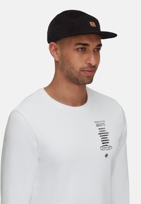 Schwarze Cordmütze mit Logo-Patch, getragen zu einem weißen Langarmshirt mit schwarzem Text und Grafiken, das ein lässiges Profil zeigt.