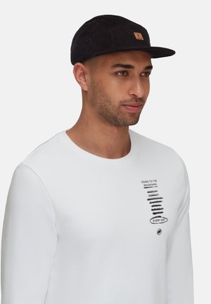 Sort fløjlskasket med logo-patch, iført en hvid langærmet skjorte med sort tekst og grafik, der viser en afslappet profil.