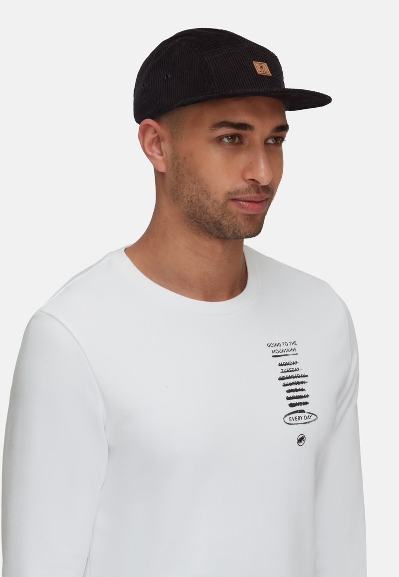 Schwarze Cordmütze mit Logo-Patch, getragen zu einem weißen Langarmshirt mit schwarzem Text und Grafiken, das ein lässiges Profil zeigt.