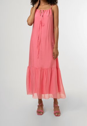 Femme portant une robe maxi rose fluide sans manches avec des bretelles fines et un détail noué au niveau de l'encolure, associée à des sandales roses à brides.