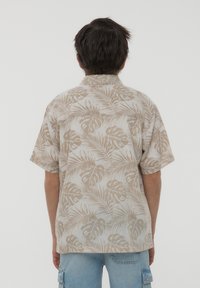 Camicetta a maniche corte in tessuto beige, con motivo a foglie di palma. Vestibilità comoda con colletto e dettagli di cucitura sulla spalla.