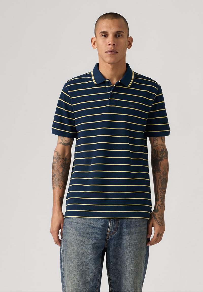 Mand med tatoveringer iført navyblå polo med gule vandrette striber og blå jeans, stående mod en ensfarvet lys baggrund.