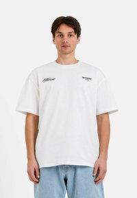 Camiseta de algodón blanca con un ajuste relajado, que presenta impresiones "Moment" y "SIXTHJUNE PARIS" en negro, mangas cortas y cuello redondo.