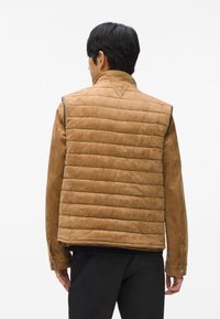 Brūns dūnu vests ar zamšā, ar strukturētu apkakli un tievu piegriezumu. Ievērojami pelēki akcenti uz pleciem un poga pie rokām.