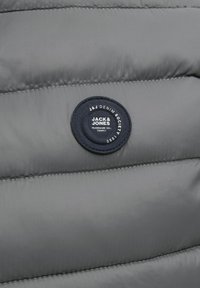 Pelēka, mīksta auduma ar quilted tekstūru, kurā ir kārtaina pagaidām zila uzlīme ar uzrakstu "JACK & JONES", kas izšūta ar baltām teksta detaļām.