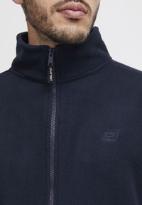 Chaqueta de forro polar en color navy con cuello alto y cremallera frontal. Presenta un logo sutil en el pecho y un acabado suave y texturizado.
