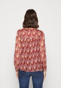 Donna con capelli castani fino alle spalle che indossa una blusa rossa a maniche lunghe con un motivo e jeans blu, girata di spalle su uno sfondo semplice.