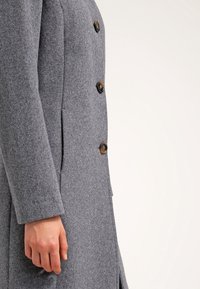 Grå ullrock med tre bruna knappar, långa ärmar, bärs av en person med en avslappnad hand vid sidan av kroppen.