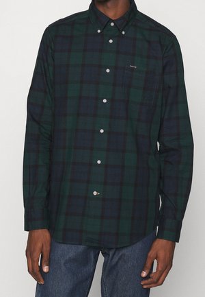 Homme portant une chemise à carreaux vert foncé et bleu marine à boutons avec une poche poitrine et un jean foncé sur un fond uni.