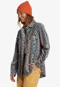 Flannel shirt with geometric patterns in blue, pink, and orange; features a buttoned front, collar, and a chest pocket. Complements beige pants.