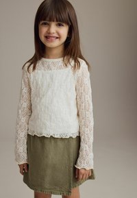 Hvid blonde langærmet top med bølget kant, parret med en grøn, knælang denimskirt. Glat tekstur med delikate blomster mønstre.
