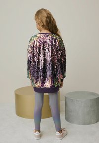 Pailletteret jakke med multikolorede reflekterende teksturer, lilla base, kombineret med gradient leggings og lyse sko. Blød baggrund.