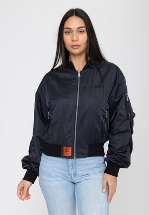 Vrouw met een zwarte bomberjack met rits, ritssluitingen op de zakken en lichtblauwe spijkerbroek, staand tegen een eenvoudige witte achtergrond.