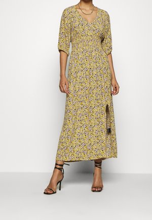 Mujer que lleva un vestido midi amarillo con estampado floral, mangas hasta el codo, abertura lateral y sandalias negras de tacón alto con tiras.