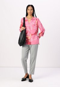 Rosa Bluse mit floralem Design, graue Hose, schwarze Handtasche und schwarze Schuhe mit strukturierter Oberfläche. Das Modell steht mit offener Körperhaltung.