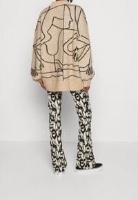 Übergröße beigefarbener Strickpullover mit schwarzem abstraktem Design, kombiniert mit schwarzen und cremefarbenen gemusterten Schlaghosen und schwarzen Sneakers.