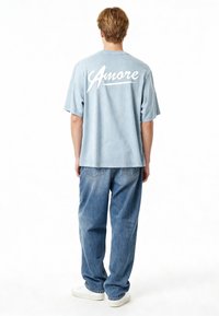 Ljusblå överdimensionerad t-shirt med vit "Amore" text på baksidan, kombinerad med löst sittande ljusblå jeans och vita sneakers.