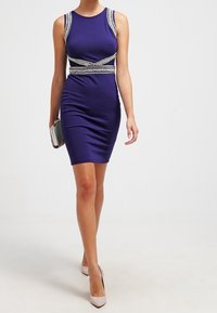 Fialové bezrukávové bodycon šaty s metalickými ozdobami na výstřihu a pase, hladká textura, doplněné stříbrnou kabelkou.