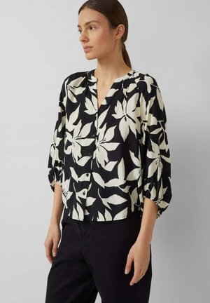 Frau trägt eine schwarze Bluse mit großen weißen Blumenmustern, dreiviertel Puffärmel und schwarze Hose, steht vor einem einfarbigen Hintergrund.