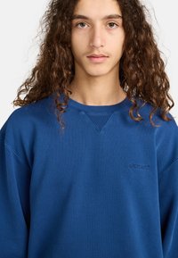 Blue sweatshirt with a ribbed neckline and a small, embossed logo on the chest. Fabric appears smooth with a relaxed fit.