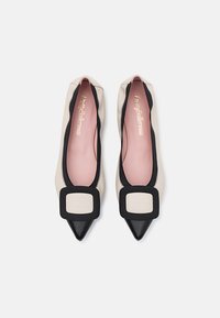 Balletskor med en krämfärgad kropp, svart tåhuvud och fyrkantig accent. Mjukt material med dragkedjedetaljer längs kanterna.