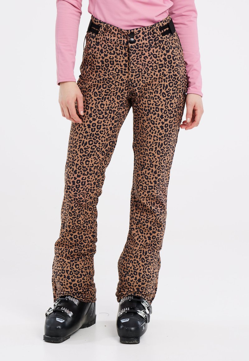 Personne portant un pantalon de ski à imprimé léopard, un haut rose à manches longues et des chaussures de ski noires, debout sur un fond blanc.
