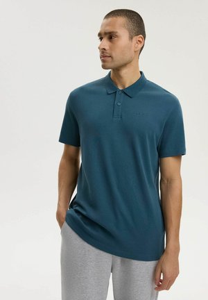 Mężczyzna w turkusowej koszulce polo z krótkim rękawem i jasnoszarych spodniach, stojący z jedną ręką w kieszeni, patrzący na bok na jednolitym tle.