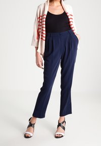 Námořnické kalhoty s vysokým pasem a upraveným střihem, z měkké tkaniny, se stranovými knoflíky. V kombinaci s krémovým kardigánem se červenými pruhy. Černo-bílé sandály.