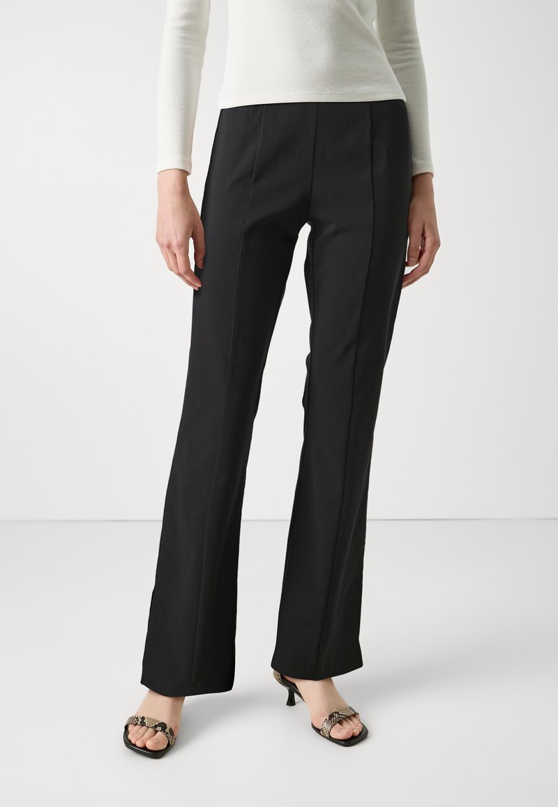 Femme portant un pantalon noir droit, un top blanc à manches longues et des sandales à talons ouvertes avec des brides imprimé serpent.