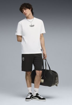 Jonge man in wit t-shirt en zwarte korte broek met een zwarte Puma weekendtas, staand tegen een neutrale grijze achtergrond.
