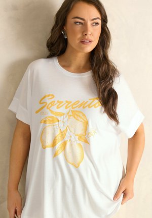 Frau mit langen braunen Haaren trägt ein weißes T-Shirt mit gelben Zitronen und dem Schriftzug "Sorrento Italy" und steht vor einem beigen Hintergrund.