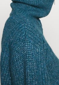 Pull en tricot de couleur bleu sarcelle foncé avec un col haut et replié. Il présente un tissage lâche texturé et de subtiles variations de ton dans le tissu.