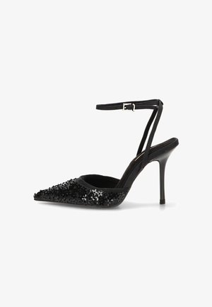 Zapato negro de tacón alto con punta puntiaguda, correa en el tobillo y lentejuelas negras decorativas en la parte delantera.