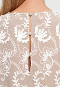 Šviesiai rudas audinys su baltais gėlių siuvinėjimais ir trimis audiniu aptrauktomis sagomis vertikaliai nugaroje esančio drabužio plyšio srityje.