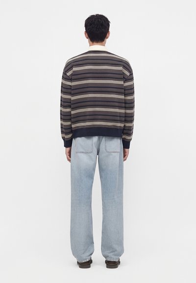 Homme portant un pull rayé foncé et un jean large bleu clair, debout de dos devant un fond uni.
