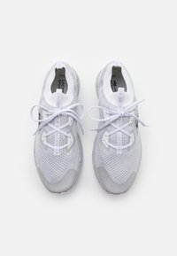 Chaussures de sport blanches avec une tige en mesh, des détails texturés et une semelle grise claire. Présentent un logo contrastant et des lacets plats.